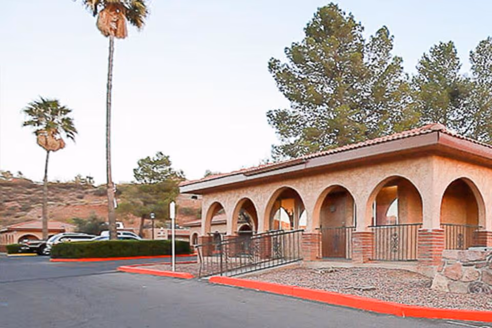 Front exterior of a stucco building with arched doorways, a ramp, palm trees, and a parking lot.