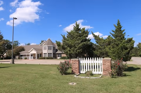 Green lawn with a white picket fence and brick posts in front of a two-story residential building with a turret under a blue sky.