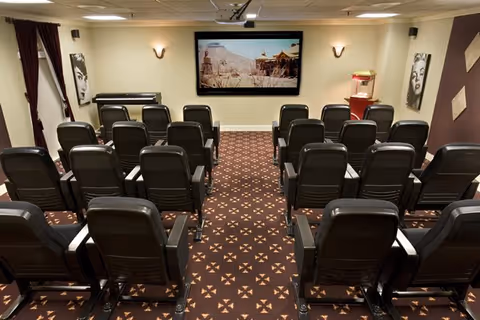 A small theater room with several rows of black leather chairs facing a large screen displaying a desert scene. The room has patterned carpet, wall sconces for lighting, a popcorn machine on the right side, and black-and-white portraits on the walls.