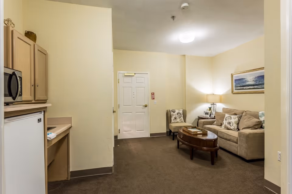 Small living area with a beige sofa, armchair, coffee table, lamp and a kitchenette near the entrance door.