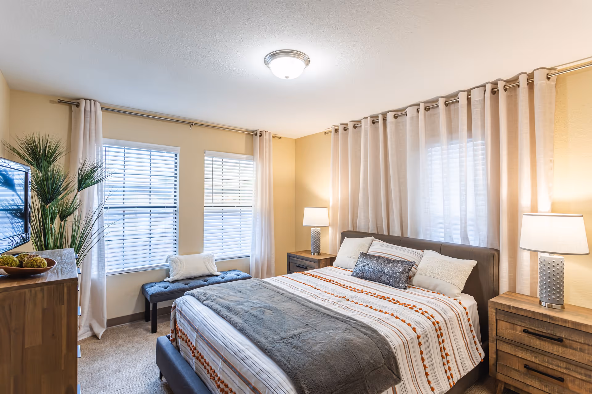 A bright and cozy bedroom with a large bed featuring striped bedding and multiple pillows. The room has two windows with white blinds and sheer curtains, two wooden nightstands with matching lamps, a wooden dresser with a TV on top, and a tall green plant in the corner. The walls are painted a soft beige color.