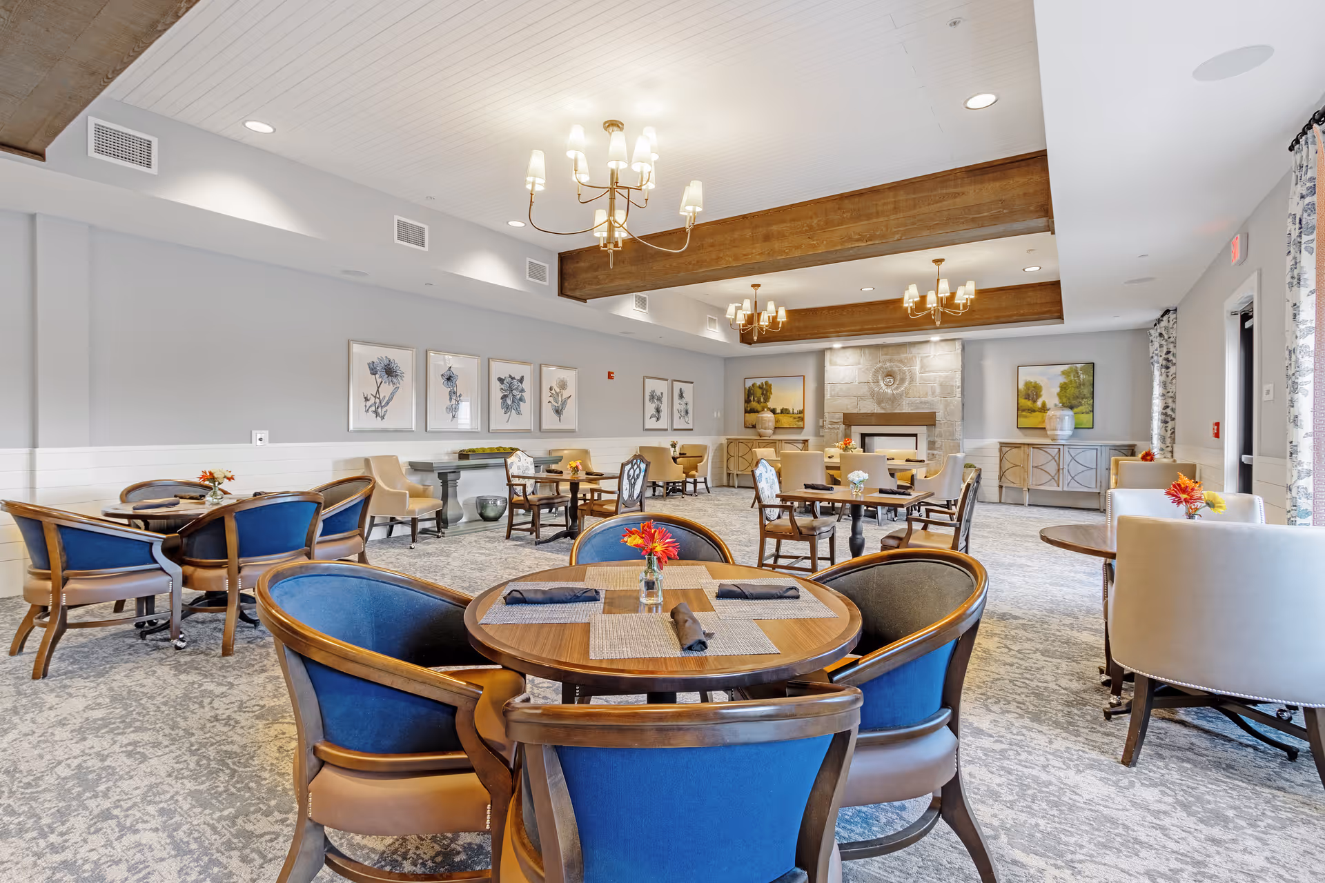 A spacious and well-lit dining room with round wooden tables and blue cushioned chairs. The room features a carpeted floor, wooden ceiling beams, chandeliers, framed floral artwork on the walls, and a stone fireplace at the far end. There are also sideboards with decorative vases and large windows with patterned curtains allowing natural light to enter.