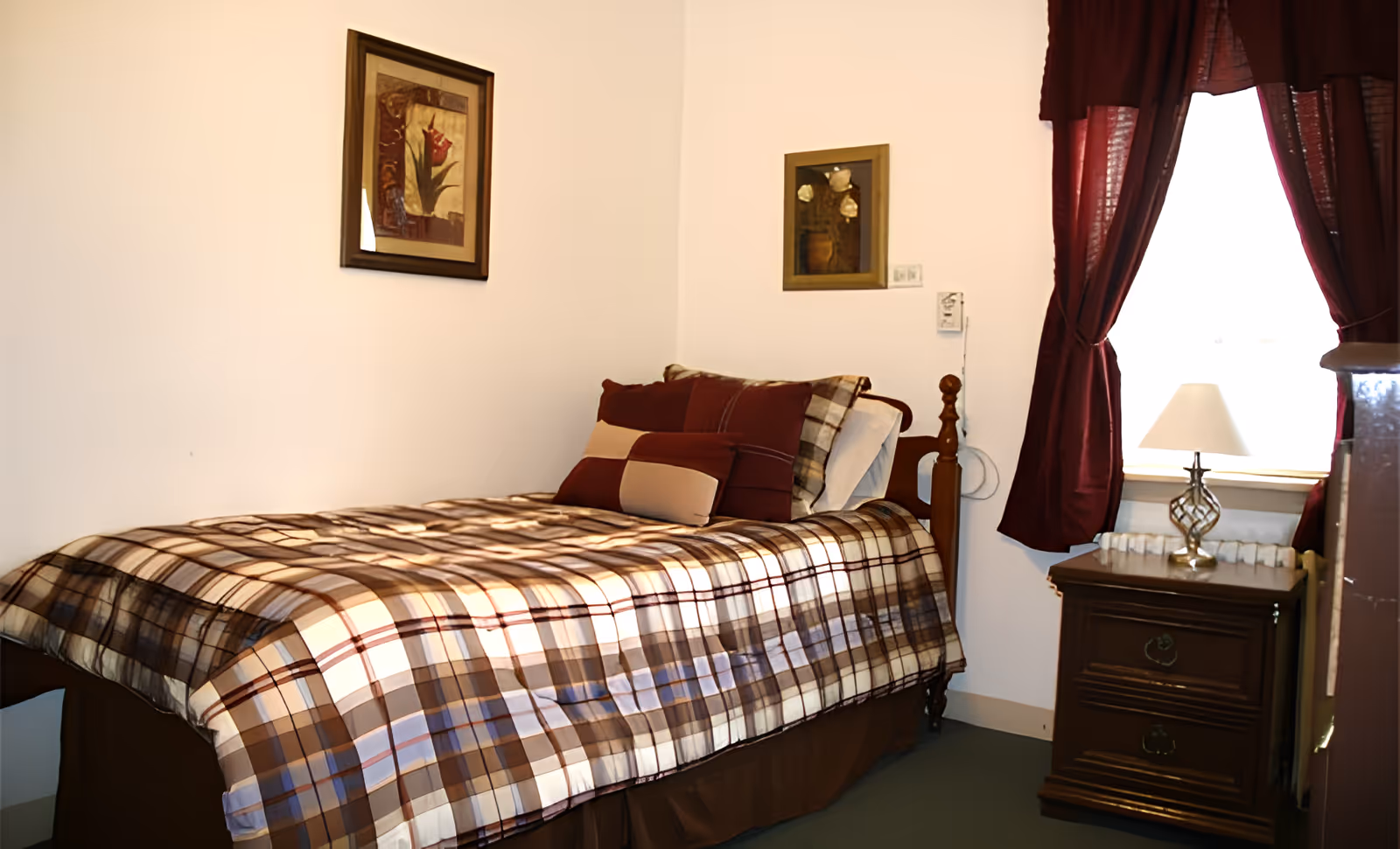 A small bedroom with a single bed covered in a brown and beige plaid bedspread and multiple pillows. There are two framed pictures on the walls above the bed. A wooden nightstand with two drawers is next to the bed, holding a decorative lamp. The window beside the nightstand is covered with dark red curtains.