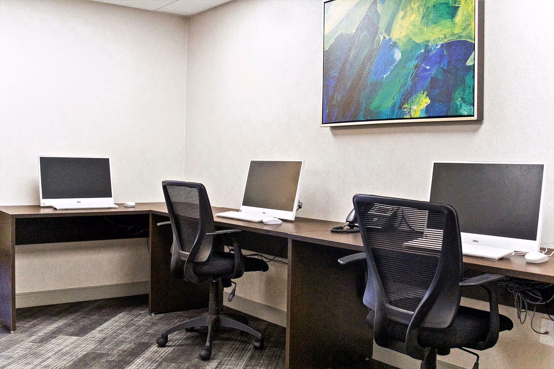 A small computer room with three desktop monitors and mesh office chairs beneath an abstract painting.