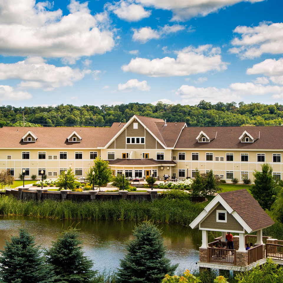 Two-story assisted living building beside a pond with a small lakeside gazebo in the foreground and trees under a partly cloudy sky.