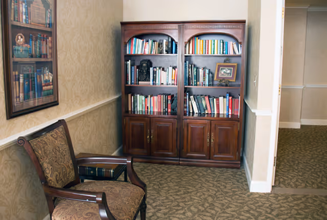 A cozy corner in an interior space featuring a wooden bookshelf filled with books and a framed picture on top. Next to the bookshelf is an upholstered wooden armchair with a patterned fabric. The walls have a beige patterned wallpaper, and there is a framed picture of books hanging on the wall to the left. The floor is carpeted with a patterned design.