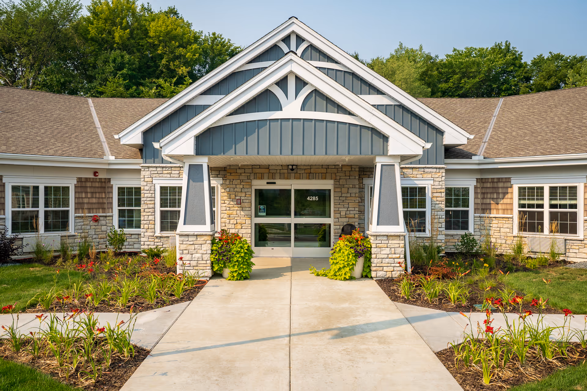 Front entrance of a single-story senior living facility with stone and wood siding, a peaked roof, and a concrete walkway leading to double glass doors. The entrance is flanked by potted plants and landscaped flower beds with green shrubs and red flowers. Trees are visible in the background.