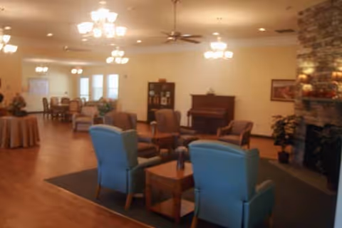 Spacious senior living common room with upholstered chairs around a coffee table, a fireplace and piano, and dining tables in the background.