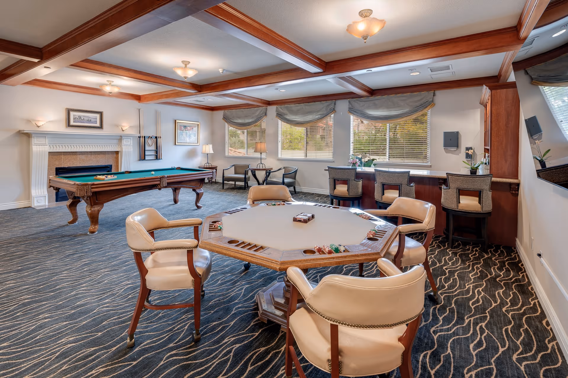 A spacious game room with a poker table surrounded by four cushioned chairs in the foreground, a pool table near a white fireplace on the left, and a bar counter with four high chairs in front of windows with blinds. The room features a patterned carpet, wooden ceiling beams, and soft lighting.