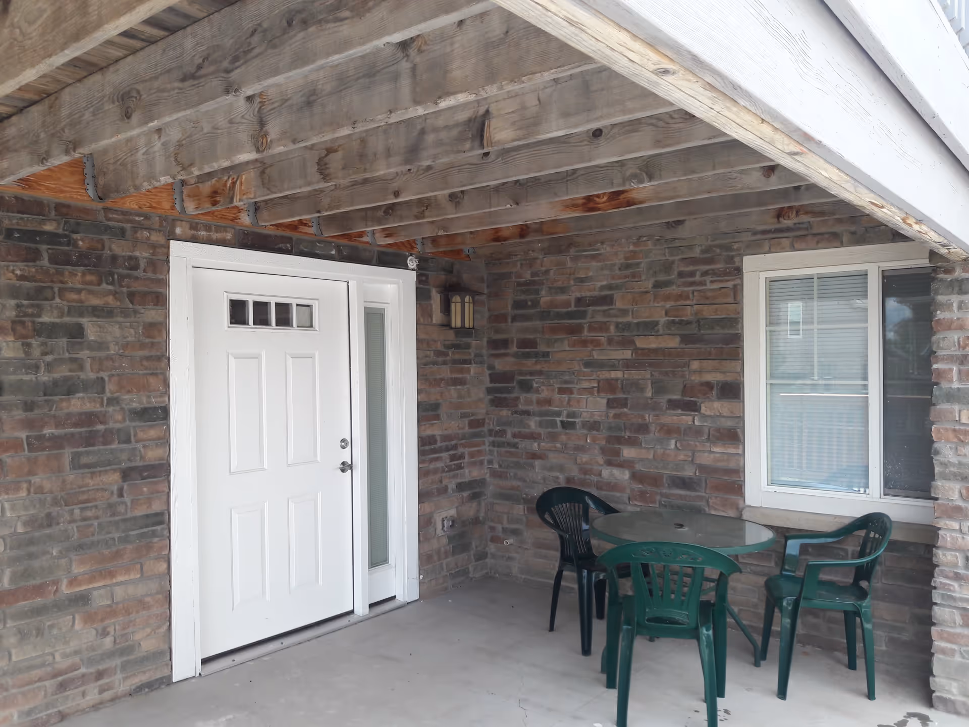 Covered outdoor patio area with a white door, a window, and a round glass table surrounded by four green plastic chairs. The walls are made of brick and the ceiling shows exposed wooden beams.