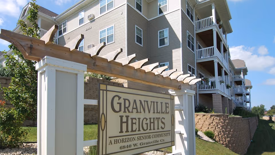 Exterior view of Granville Heights Senior Apartments showing a large sign with the community name and address in front of a multi-story residential building with balconies, under a clear blue sky.