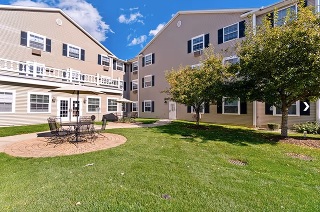 Outdoor courtyard area of a senior living facility with green grass, trees, and a round patio table with chairs. The beige multi-story building with white trim and multiple windows surrounds the courtyard under a blue sky with some clouds.