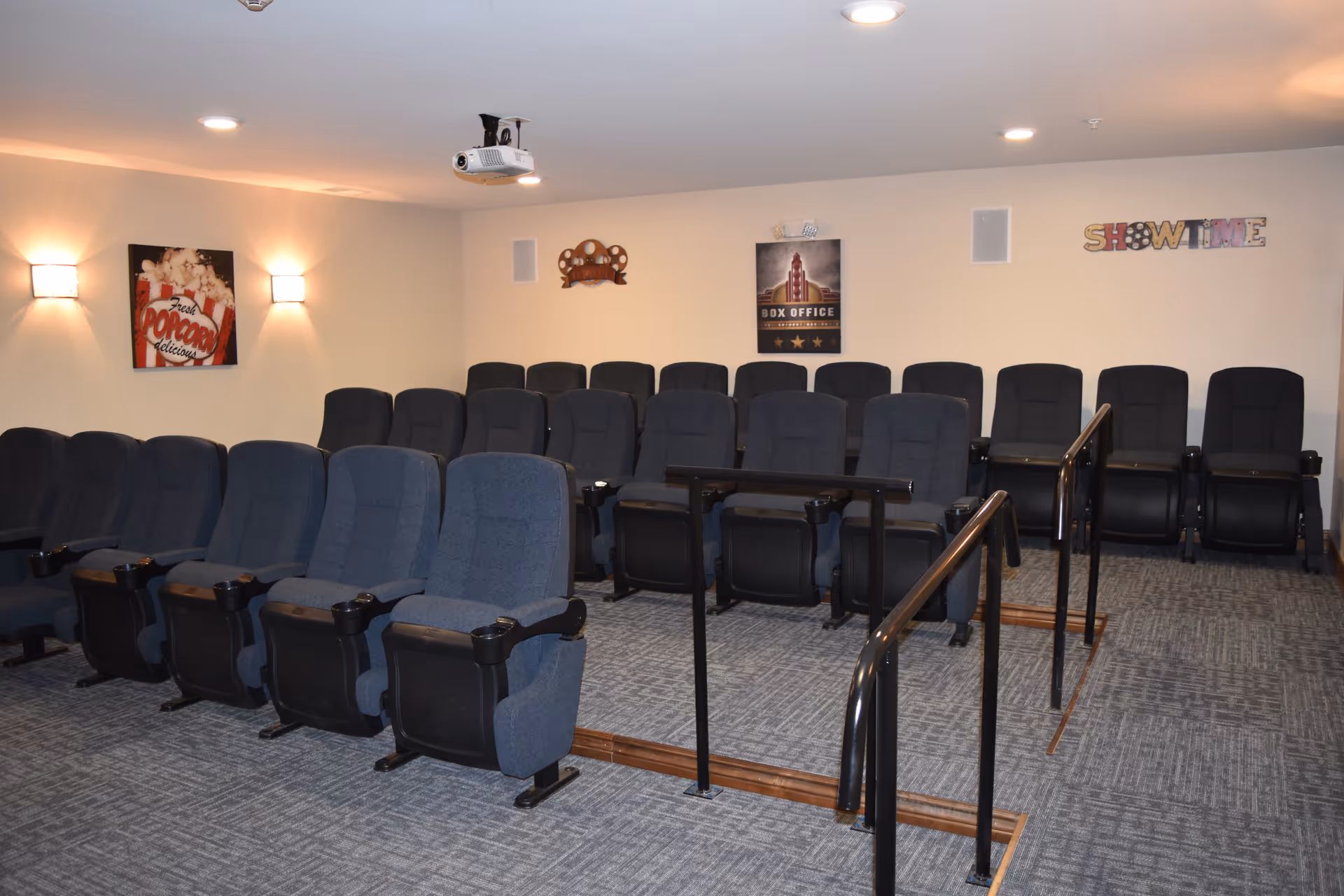 A small indoor movie theater with several rows of blue upholstered seats, a ceiling projector, and movie-themed wall decor.