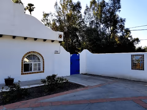 White stucco building and courtyard with an arched window and a bright blue gate in a white wall, with trees in the background.