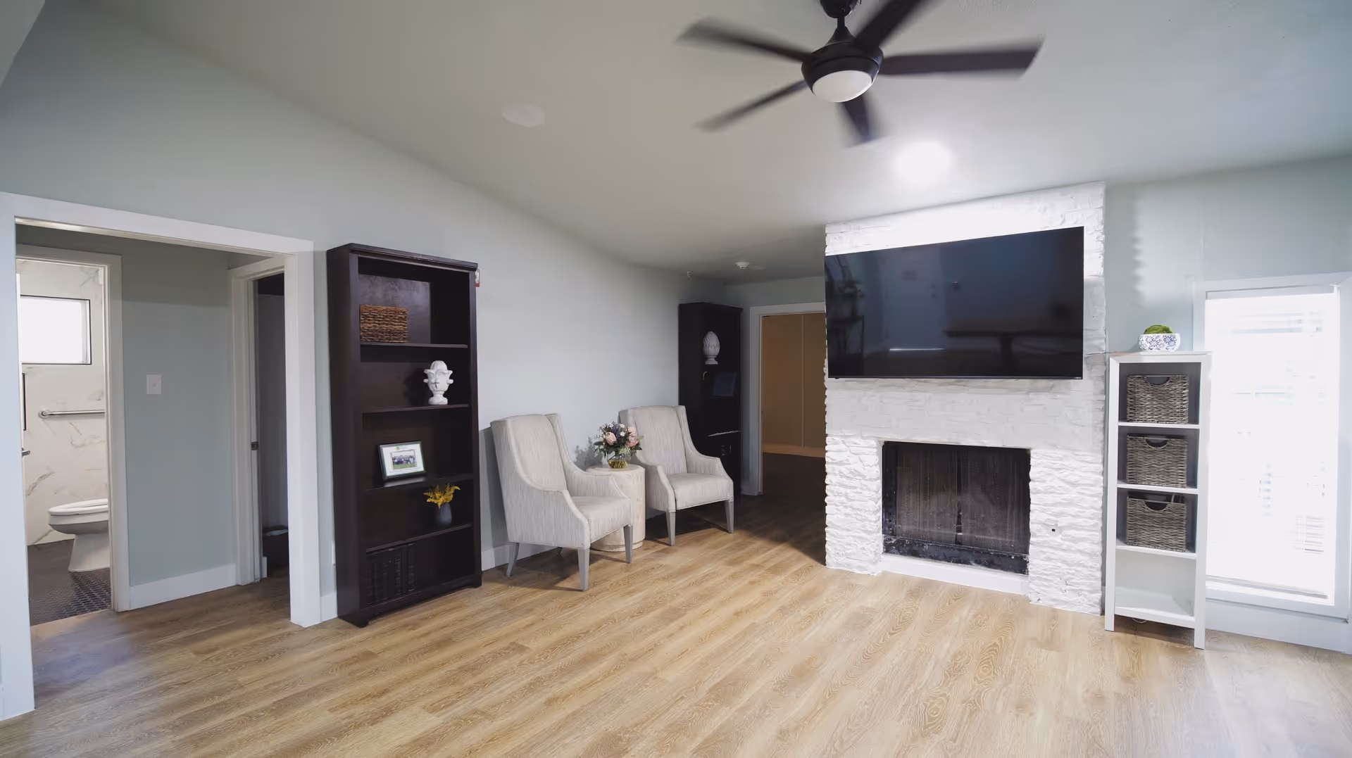 A bright living room with light wood flooring, two beige armchairs with a small table and flowers between them, a dark wooden bookshelf with decorative items, a white stone fireplace with a large flat-screen TV mounted above it, and a white shelving unit with baskets next to a window with blinds. A ceiling fan with a light is mounted on the ceiling. A doorway on the left leads to a bathroom with a visible toilet.