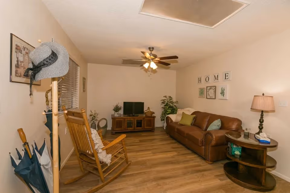 A cozy living room with a brown leather sofa adorned with green and beige pillows, a wooden rocking chair with a white throw, a wooden TV stand with a flat-screen TV, and a ceiling fan with lights. The walls are decorated with framed artwork and a set of four wall plaques spelling 'HOME'. A wooden side table with a lamp, tissues, and a book is next to the sofa. A coat rack with hats and umbrellas stands near a window with blinds.