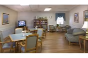 A cozy common area in a senior living facility featuring wooden floors, several tables with chairs, a television on a stand, bookshelves filled with books, framed artwork on the walls, and comfortable seating including armchairs and a loveseat near a window with a valance.
