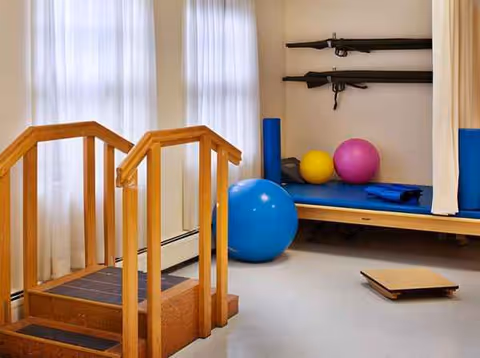 A rehabilitation room with wooden stairs and handrails, a blue exercise ball, and colorful therapy balls.