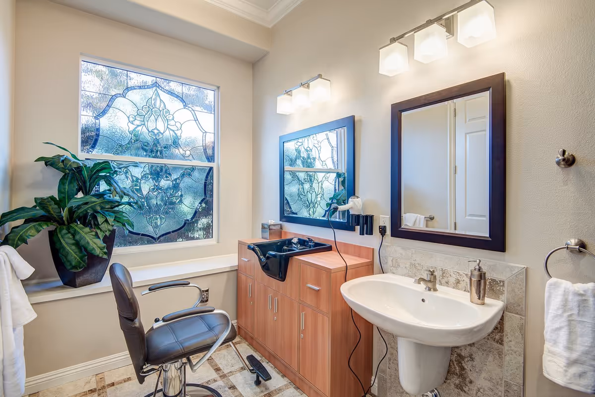 A bright and clean salon area within a facility, featuring a black salon chair in front of a wooden cabinet with a black sink for hair washing. There is a large decorative frosted window with a floral pattern, a potted plant on the windowsill, a white pedestal sink with a soap dispenser, two mirrors with dark wooden frames, and modern light fixtures above the mirrors. White towels hang on towel rings on the walls.