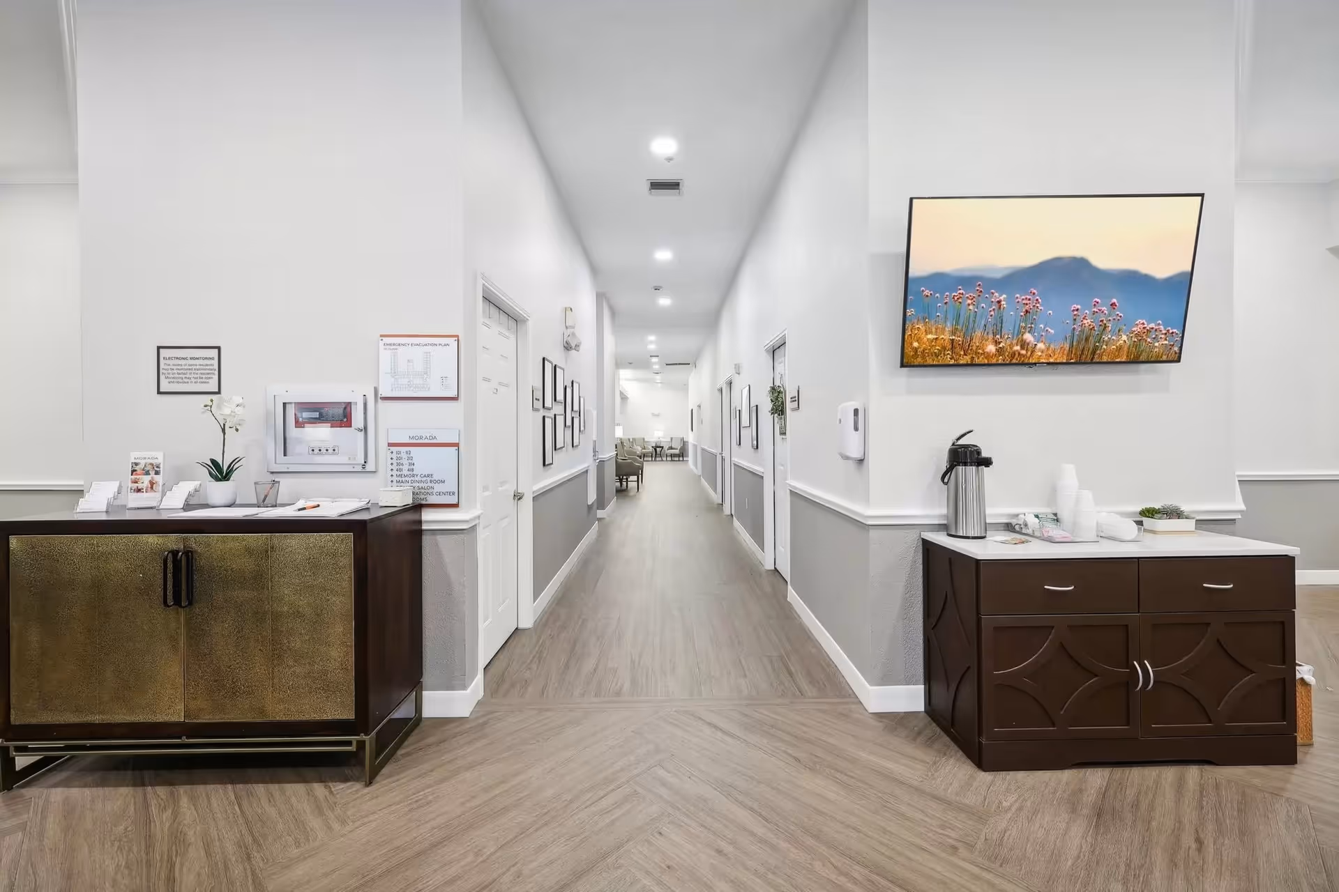 A clean, well-lit hallway in a senior living facility with light wood flooring and white walls. On the left side, there is a dark wooden cabinet with a potted orchid and informational brochures. On the right side, a dark wooden counter holds a coffee dispenser, cups, and a small plant. A flat-screen TV mounted on the wall displays an image of flowers with mountains in the background. The hallway extends into a seating area with chairs visible in the distance.