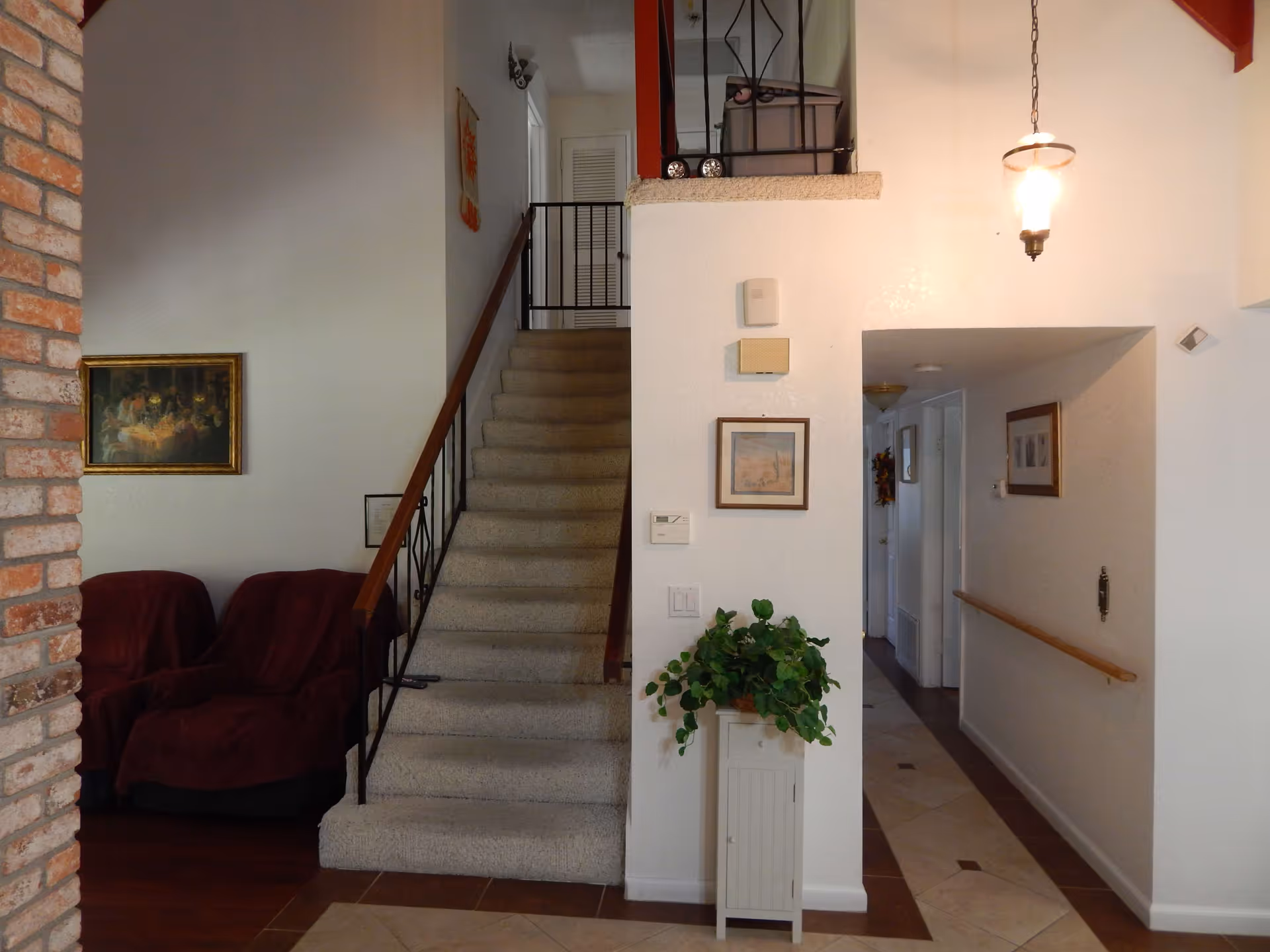 Interior view of a senior living facility showing a carpeted staircase with a wooden handrail, two armchairs covered with maroon fabric to the left, a brick wall partially visible, a hallway with framed pictures on the walls, a small cabinet with a green potted plant, and a hanging light fixture above.