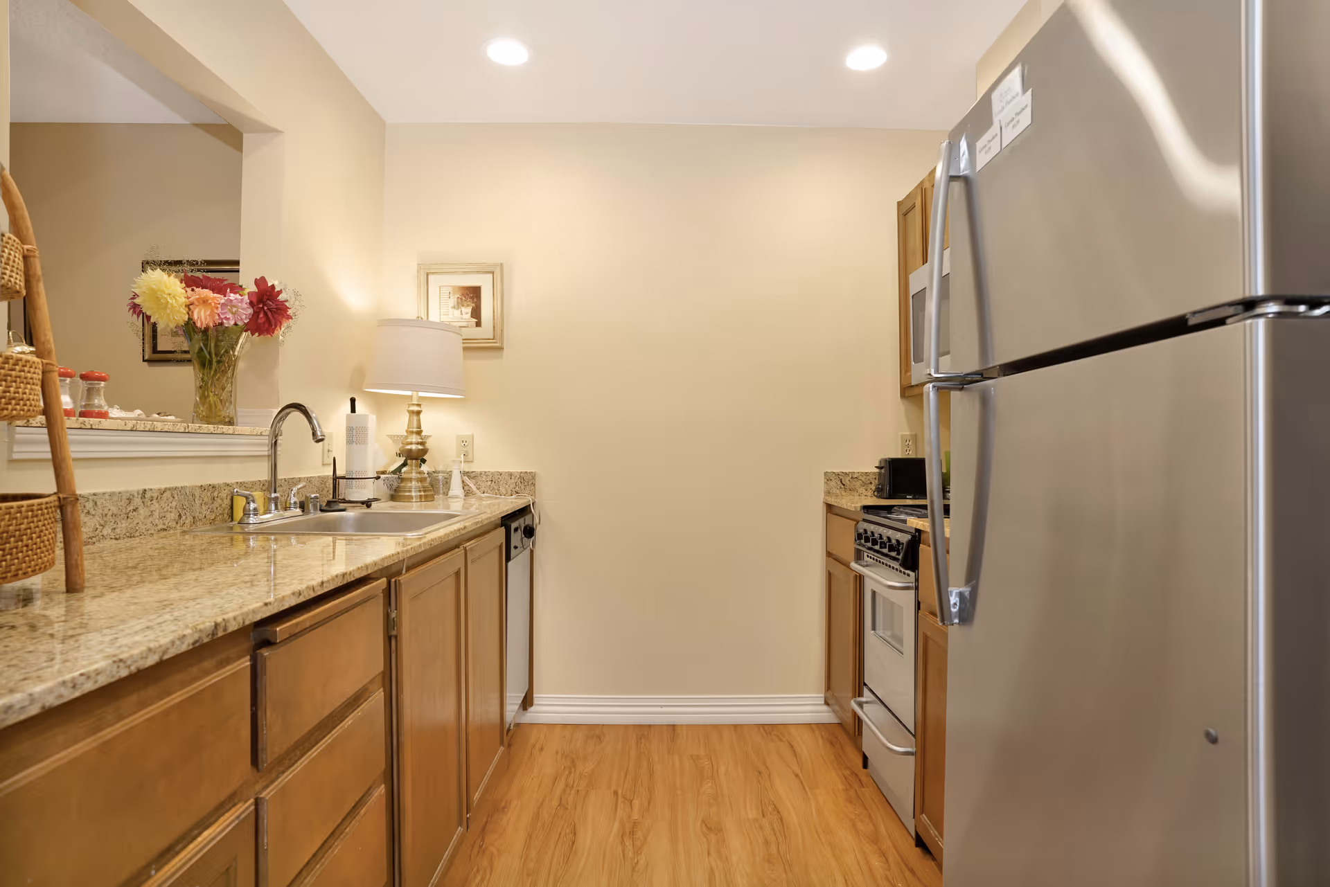 A small kitchen with wooden cabinets, granite countertops, a stainless steel refrigerator, a stove, a dishwasher, and a sink. There is a lamp and a vase with flowers on the counter, and a framed picture on the wall.