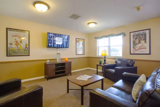 A cozy living room with beige walls and carpeted floor, featuring a dark brown leather sofa and armchairs arranged around a wooden coffee table. A flat-screen TV is mounted on the wall above a wooden media console. The room is decorated with framed vintage movie posters and has a window with a light blue valance letting in natural light. A table lamp and small plant sit on a side table next to one of the armchairs.