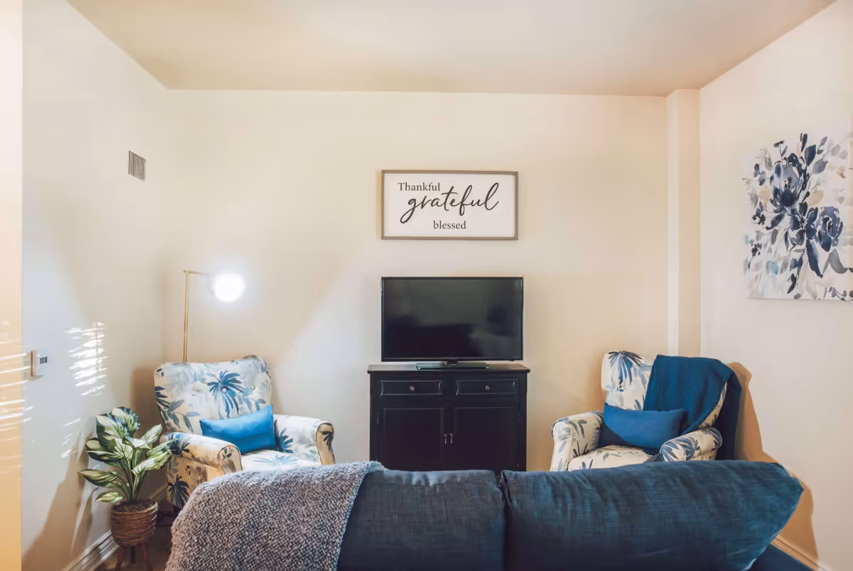 A cozy living room with two floral-patterned armchairs each with a blue cushion, a dark wooden TV stand with a flat-screen TV on top, a blue sofa with a knitted throw blanket, a potted plant on the left, and wall art including a framed sign that reads 'Thankful grateful blessed' and a floral painting.