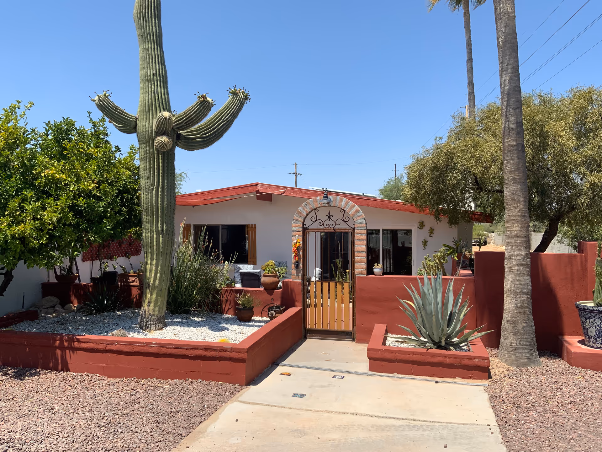 Front exterior view of a single-story building with a red roof and white walls, surrounded by desert landscaping including a large cactus, palm trees, and other plants. A gated entrance with a brick archway leads to the building.