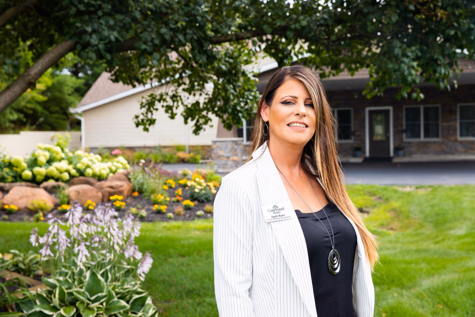 A woman with long hair wearing a white striped blazer and black top stands outside in front of a landscaped garden with flowers, rocks, and green grass. Behind her is a building with stone and siding exterior and a covered entrance. The woman is smiling and wearing a name tag that reads 'Courtyard Manor April Kern'.