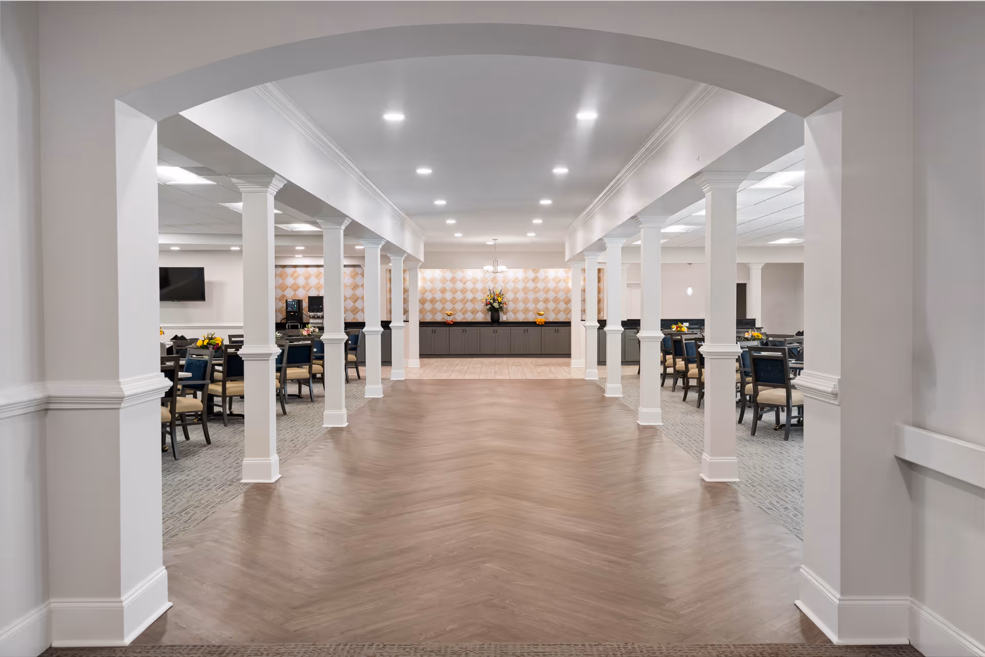 Spacious dining area in a senior living facility with multiple tables and chairs arranged on both sides of a wide hallway. The room features white columns, recessed ceiling lights, and a decorative wall with a flower arrangement at the far end.