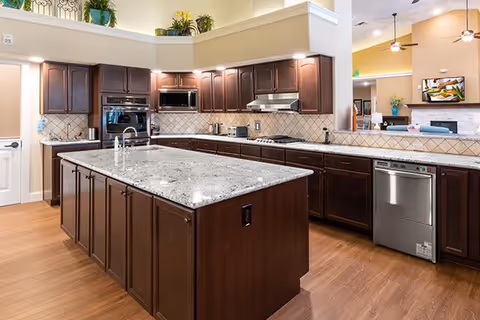 Spacious kitchen with dark wood cabinets, a large granite island with a sink, stainless steel appliances including a dishwasher, microwave, and oven, and a tiled backsplash. The kitchen opens to a living area with a ceiling fan and a mounted TV above a fireplace.