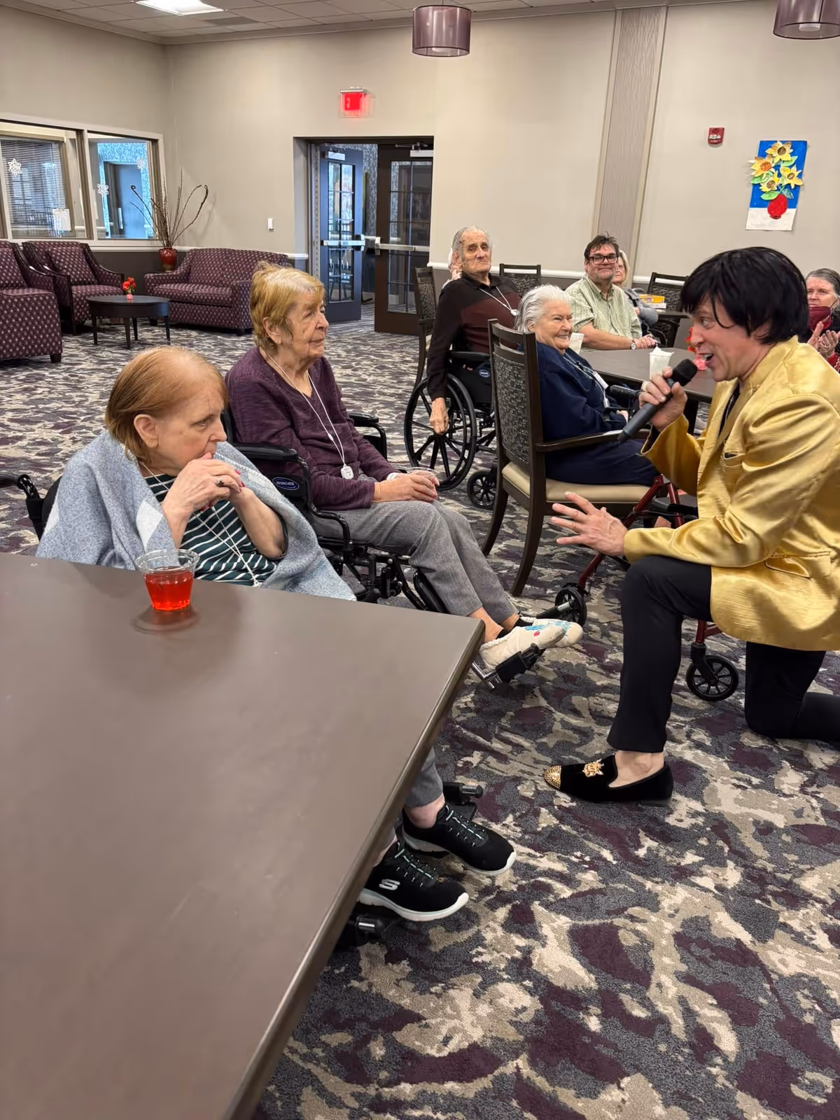 A group of elderly residents, some in wheelchairs, seated around tables in a common area. A man in a gold jacket is kneeling and singing into a microphone, engaging with the residents. The room has patterned carpet, beige walls, and purple upholstered chairs in the background.