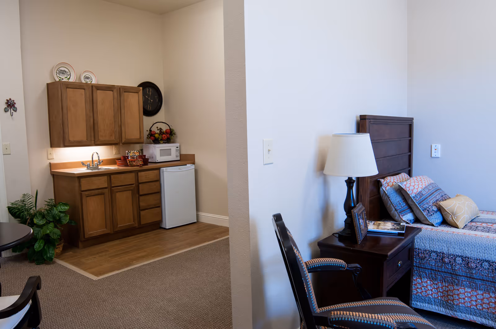 A small living space featuring a kitchenette with wooden cabinets, a sink, a microwave, and a mini refrigerator on the left side. On the right side, there is a bed with a patterned quilt, a nightstand with a lamp, a framed photo, and a book. A cushioned chair is positioned near the nightstand. The room has beige walls and carpeted flooring with a small section of wood flooring in the kitchenette area.