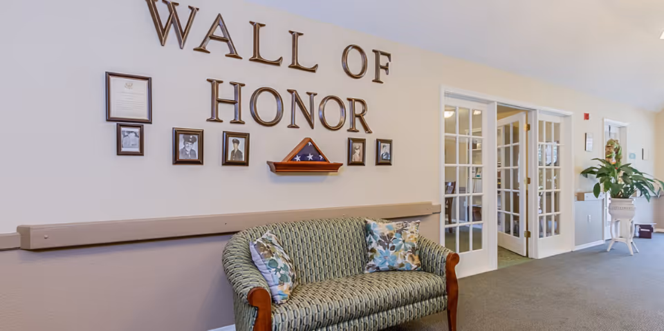 Interior hallway of a senior living facility with a green patterned couch beneath a 'Wall of Honor' display featuring framed photos and a folded American flag. To the right, there are glass-paneled double doors and a large potted plant on a white stand.