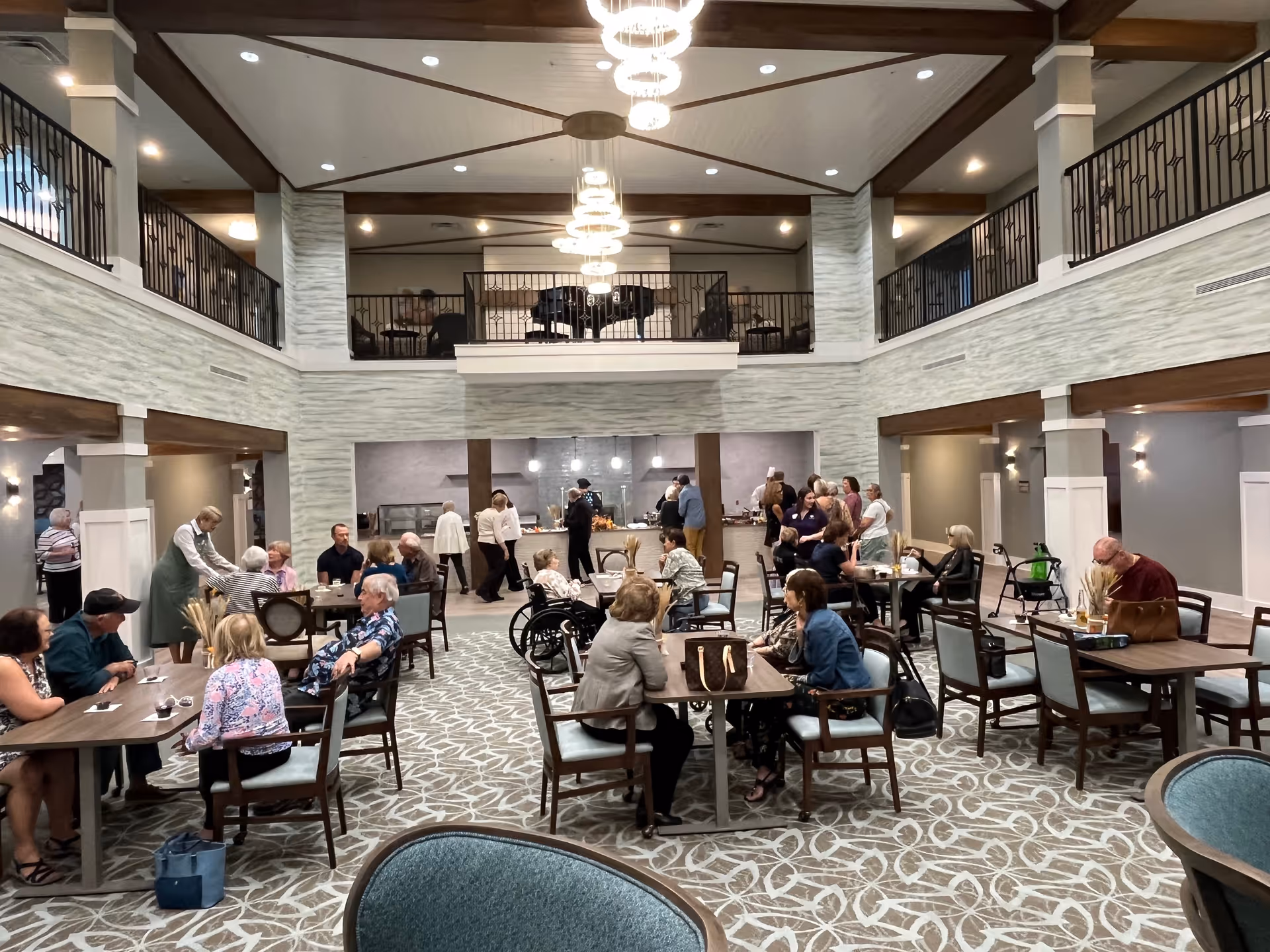 A spacious, well-lit dining area in a senior living facility with multiple tables and chairs occupied by elderly residents and staff. The room features a high ceiling with modern chandeliers, a second-floor balcony with a piano, and a serving area where people are gathered. The decor includes light-colored walls and patterned carpet flooring.