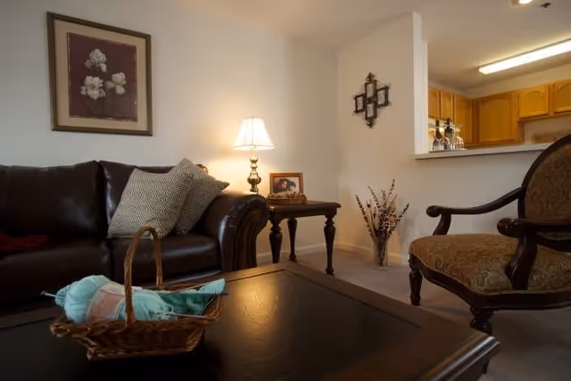 A cozy living room with a dark leather sofa adorned with patterned pillows, a wooden coffee table with a basket containing yarn and knitting needles, an upholstered armchair, a side table with a lamp and framed photo, and a partial view of a kitchen area with wooden cabinets.