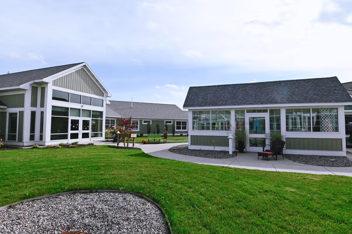 Outdoor view of The Cortland Holland Meadows facility showing green lawns, a curved concrete pathway, and two light green buildings with white trim and large windows under a partly cloudy sky.