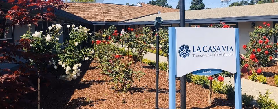 Landscaped front courtyard with rose bushes and a freestanding sign for La Casa Via Transitional Care Center in front of a single-story building.