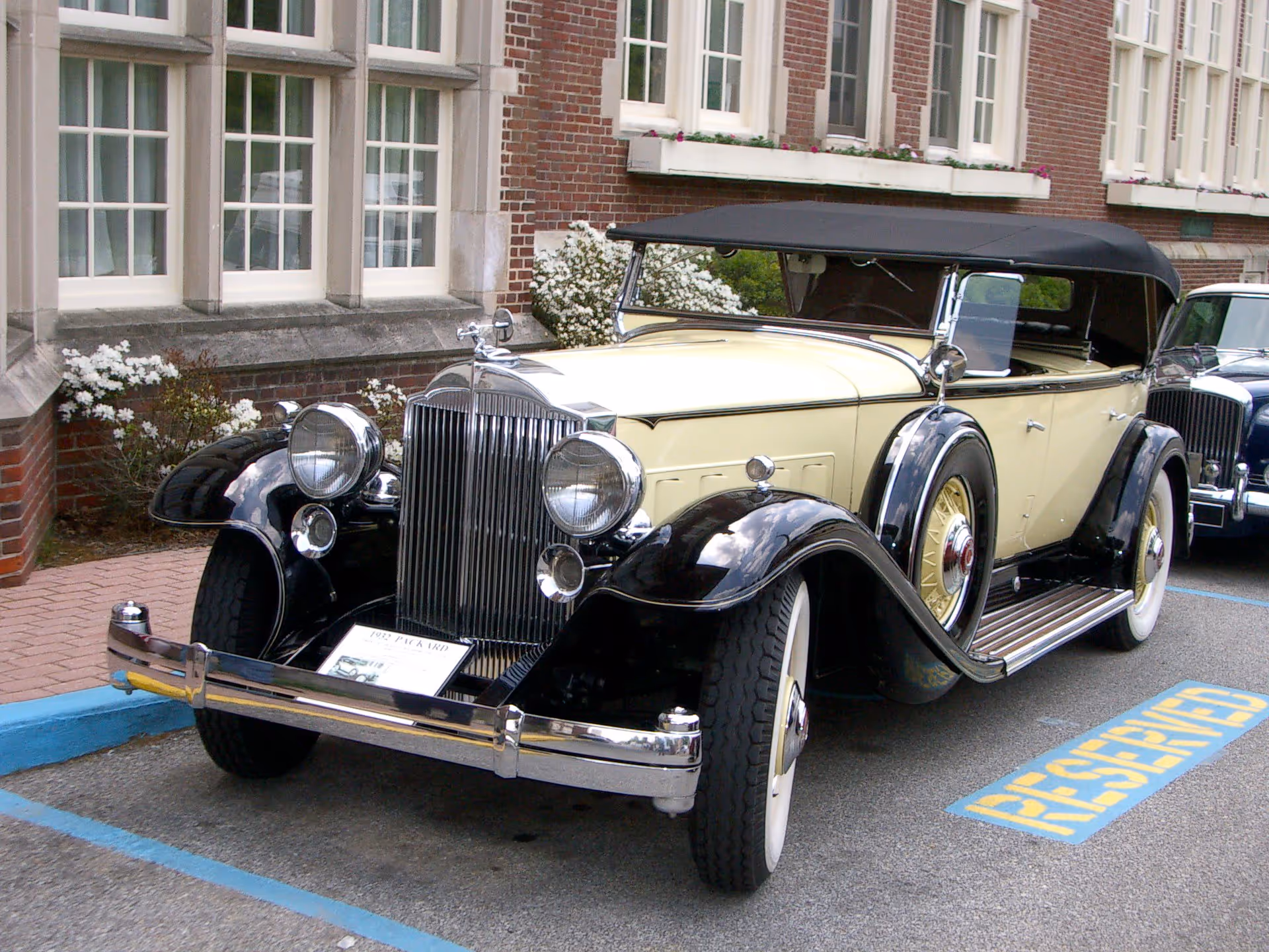A vintage cream and black convertible car parked in a reserved parking space in front of a brick building with large windows and flower boxes.