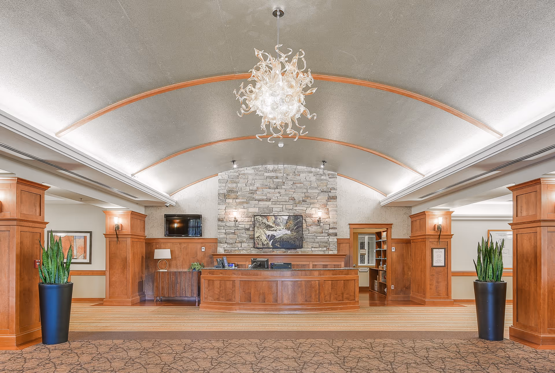 Spacious reception lobby with a curved ceiling and chandelier, central wooden front desk against a stone accent wall and large potted plants.