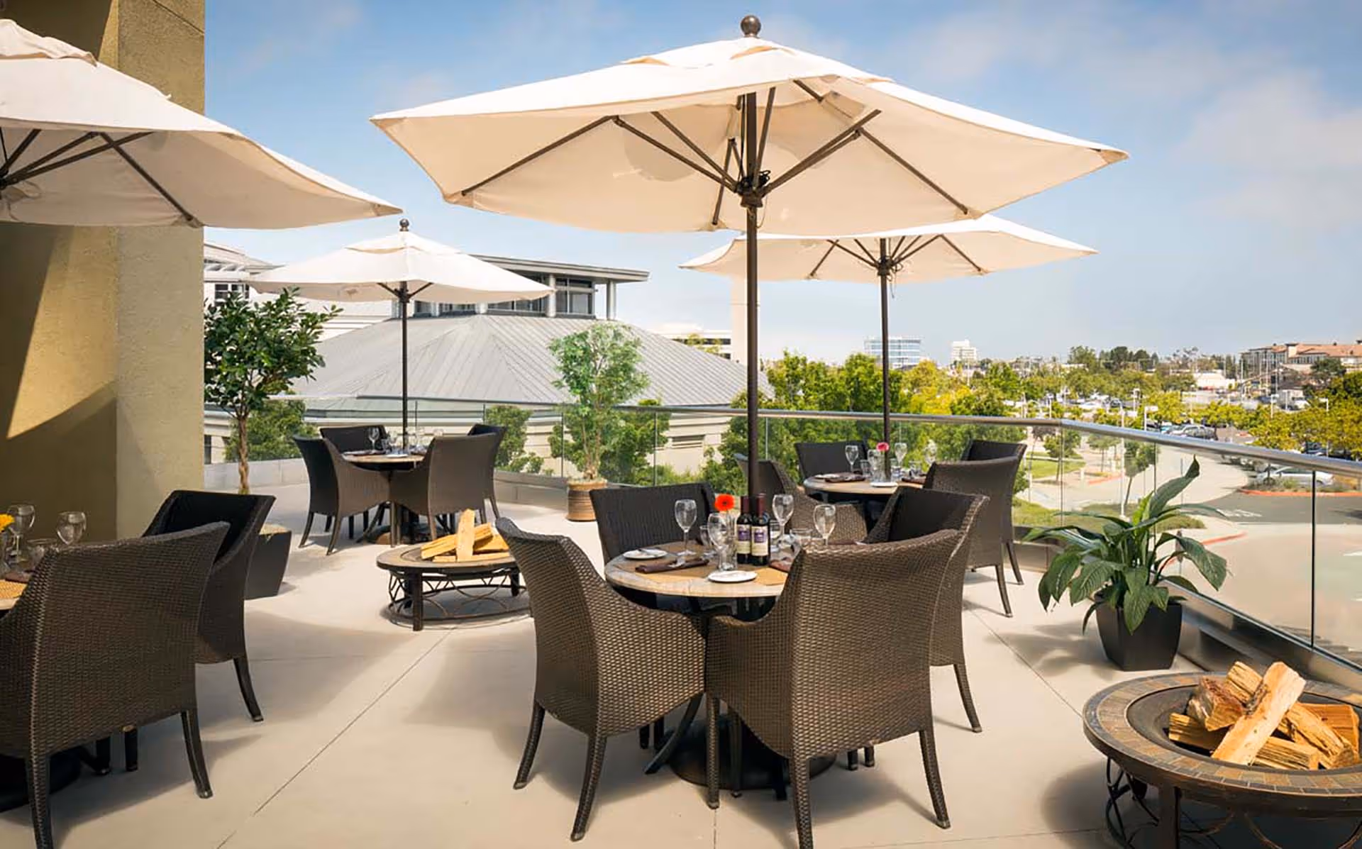Outdoor patio area with several round tables surrounded by wicker chairs, each table set with wine glasses, plates, and a bottle of wine. Large white umbrellas provide shade, and there are potted plants and a fire pit with stacked firewood. The background shows a view of nearby buildings and greenery under a partly cloudy sky.