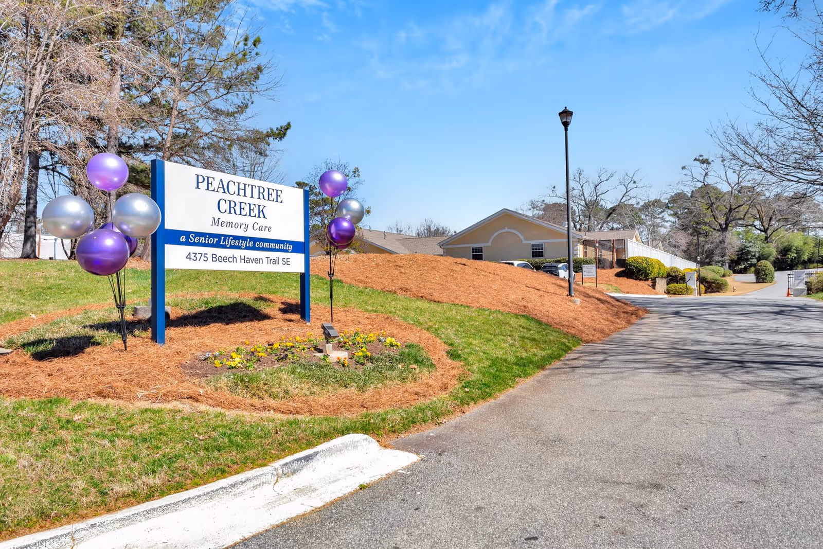 Outdoor view of Peachtree Creek Memory Care facility sign with purple and silver balloons on a grassy area next to a paved driveway. The building and parking area are visible in the background under a clear blue sky.
