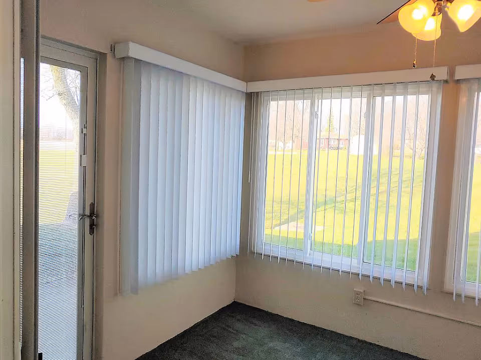 Small sunroom corner with vertical blinds on large windows and a glass door overlooking a grassy yard.