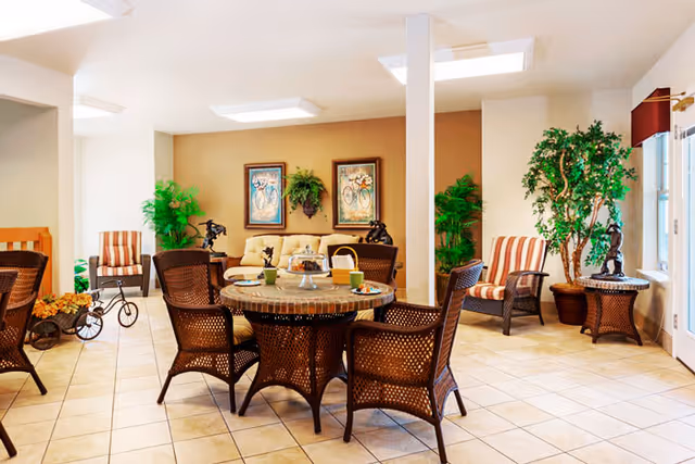 Bright common lounge with a round wicker table and chairs, sofa and armchairs, potted plants, and wall art.