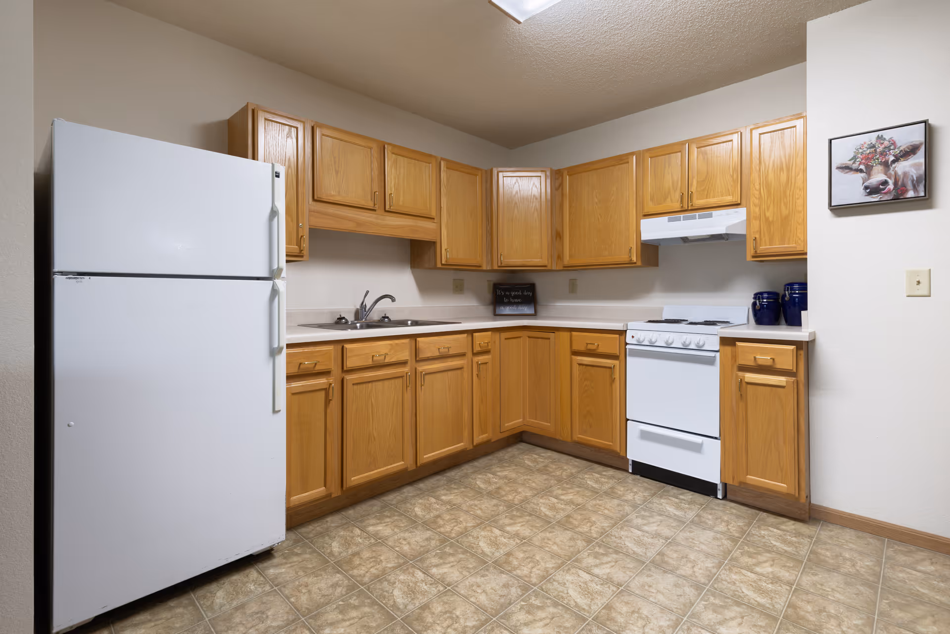 A kitchen with wooden cabinets, a white refrigerator, a white stove with an overhead vent, a double sink, and a tiled floor. There is a framed picture of a cow on the wall and a small decorative sign on the countertop.