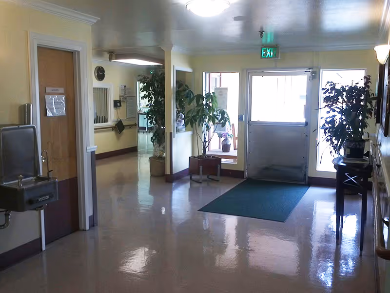 Well-lit building entrance lobby with a glass door, exit sign, potted plants, seating, and a water fountain.