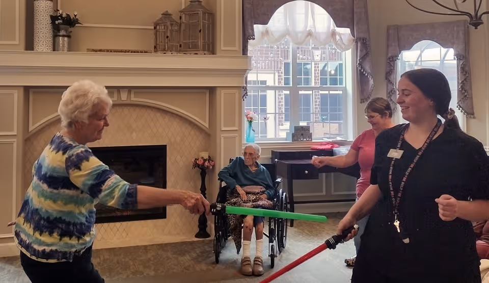 Two women, one elderly and one staff member, playfully spar with foam lightsabers in a senior living facility common area. Another elderly woman in a wheelchair and a third woman watch and smile. The room has a fireplace, large windows with curtains, and decorative items on the mantel.