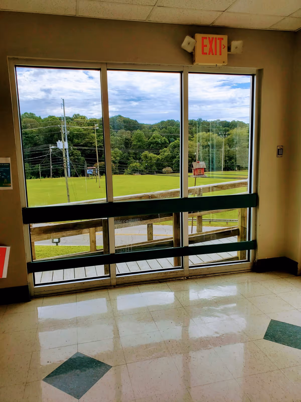 Interior view of a glass exit door with an EXIT sign overlooking a grassy lawn and trees outside.