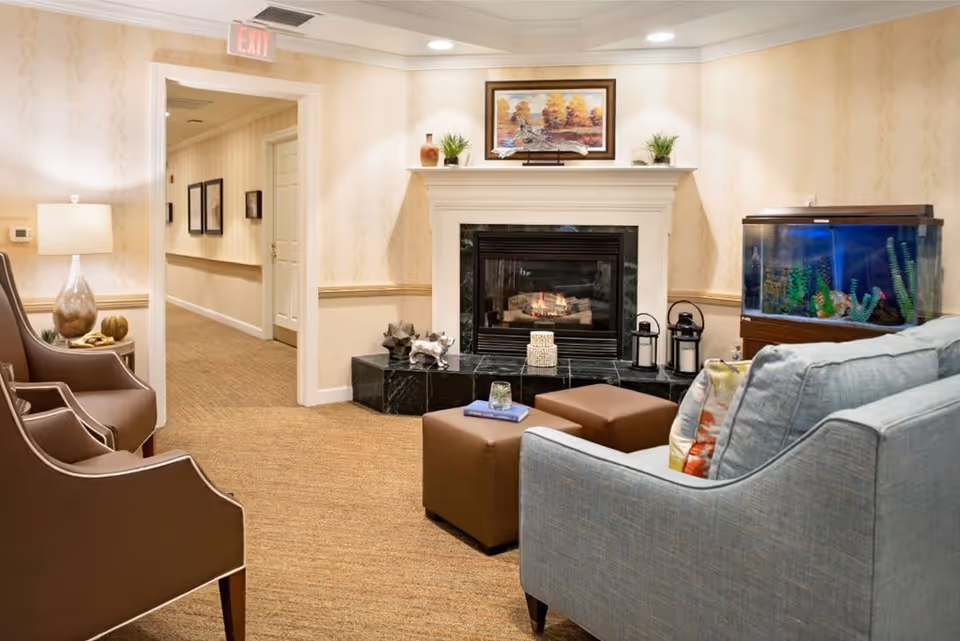 A cozy living room area in a senior living facility featuring a lit fireplace with decorative items on the mantel, a fish tank on a wooden stand, a light blue armchair with colorful pillows, two brown ottomans, and two brown chairs. The walls are decorated with wallpaper and framed artwork, and there is a hallway visible in the background with additional framed pictures.